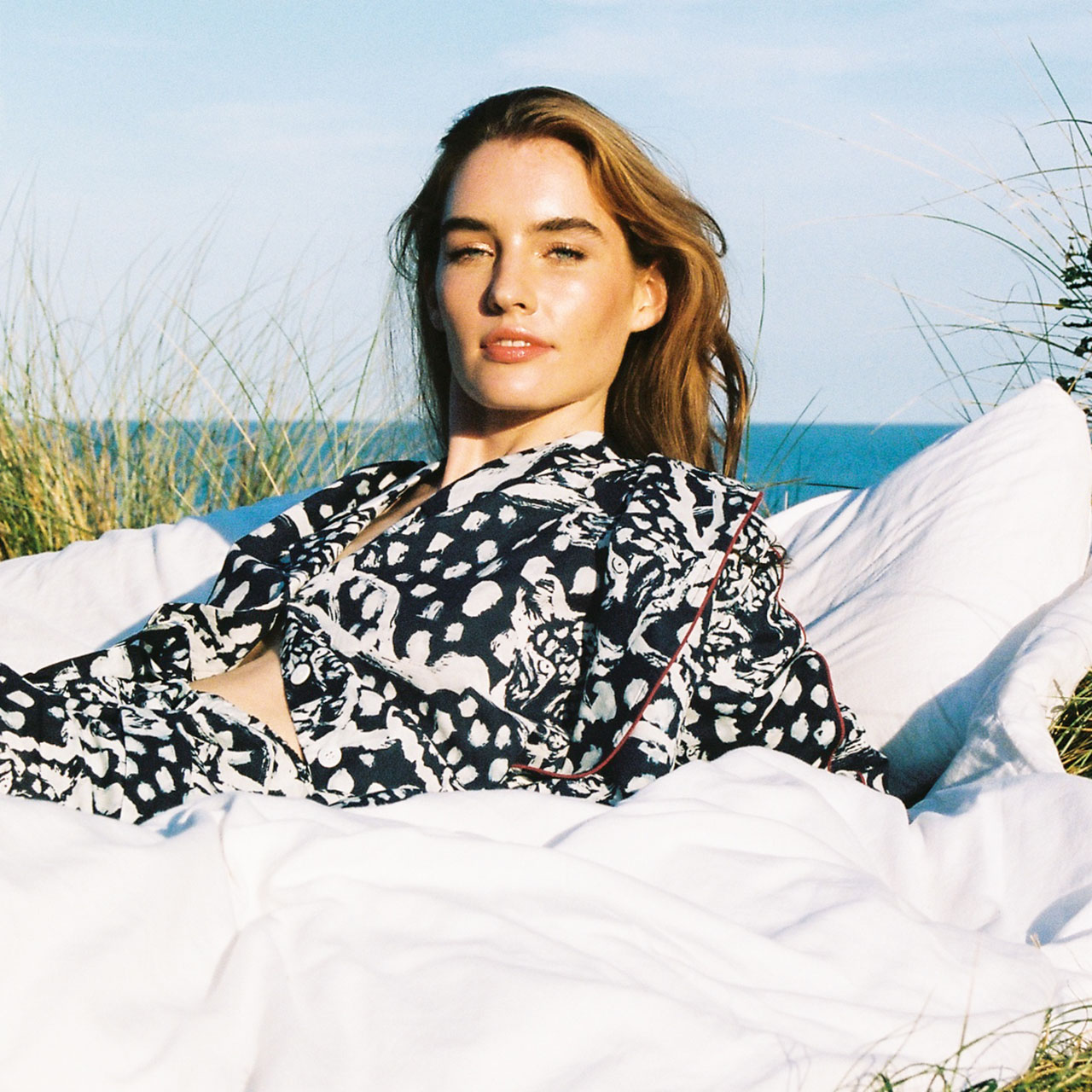 Model in coordinated printed blouse and skirt, lying on the grass in the sun, by the sea