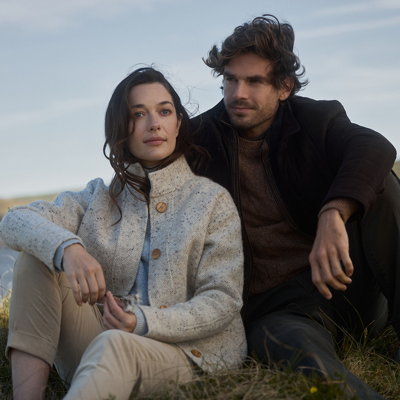 Mens and womenswear models wearing Magee coats and knit jumpers, sitting on the grass in the countryside