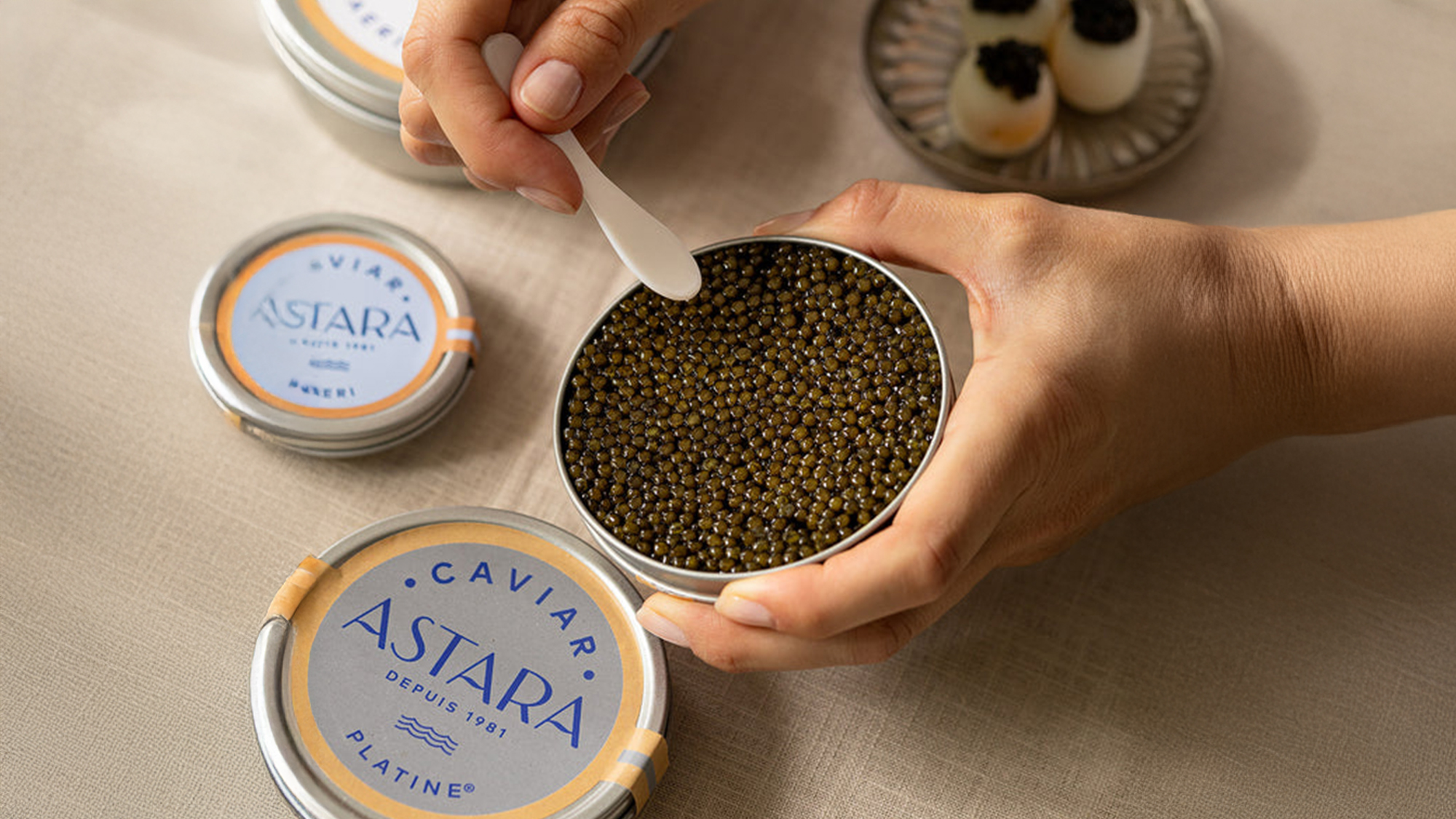 Hands holding a tin of black caviar with a small spoon, on a table with other caviar tins and caviar-topped appetizers.
