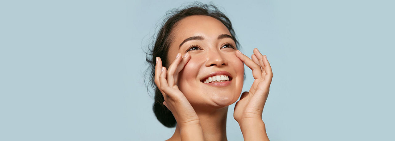 A smiling person looks upward, gently touching their face with both hands, set against a light blue background.

