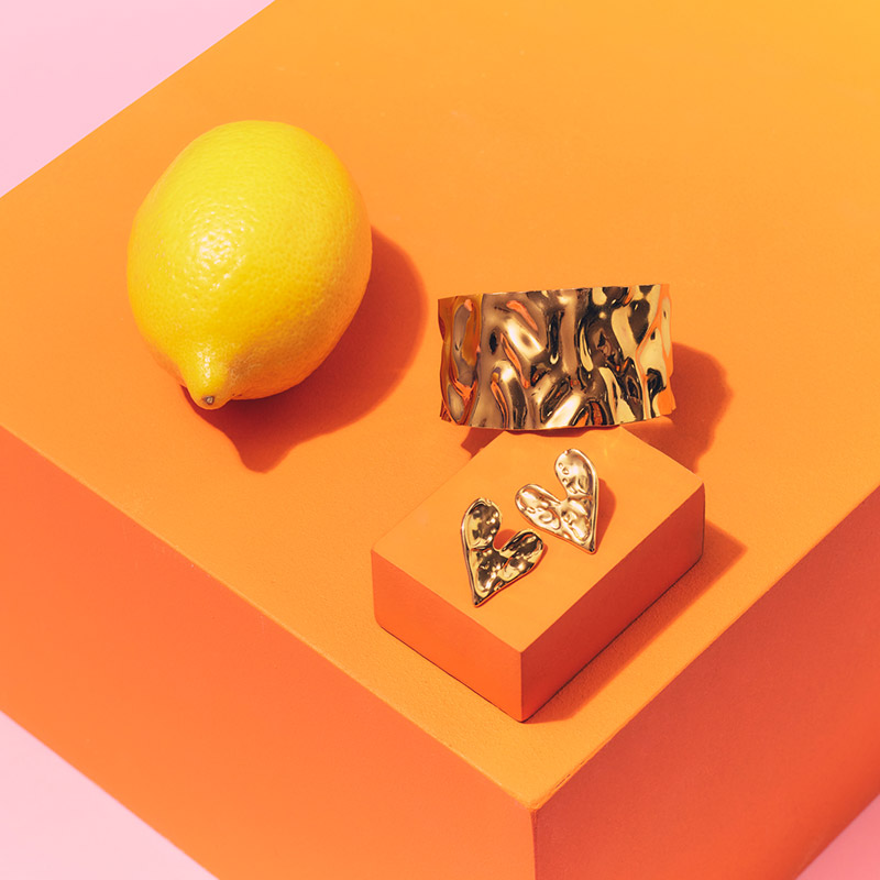 A lemon, textured gold jewelry, and an orange block are arranged against a pink background.