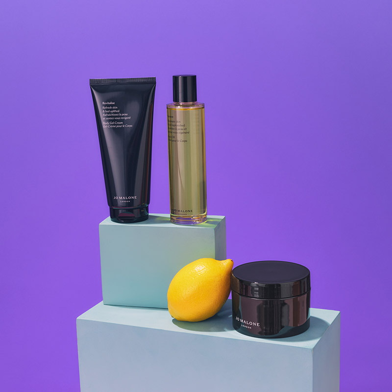 Skincare products and a lemon on pastel blocks against a purple background.