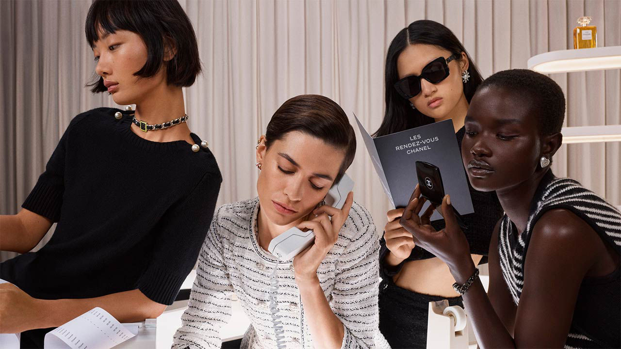 Four stylish individuals are seated indoors; one holds a phone, another reads, and two appear focused on their activities.