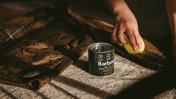 Pot of Barbour jacket wax in foreground, with someone waxing brown Barbour jacket in background with sponge