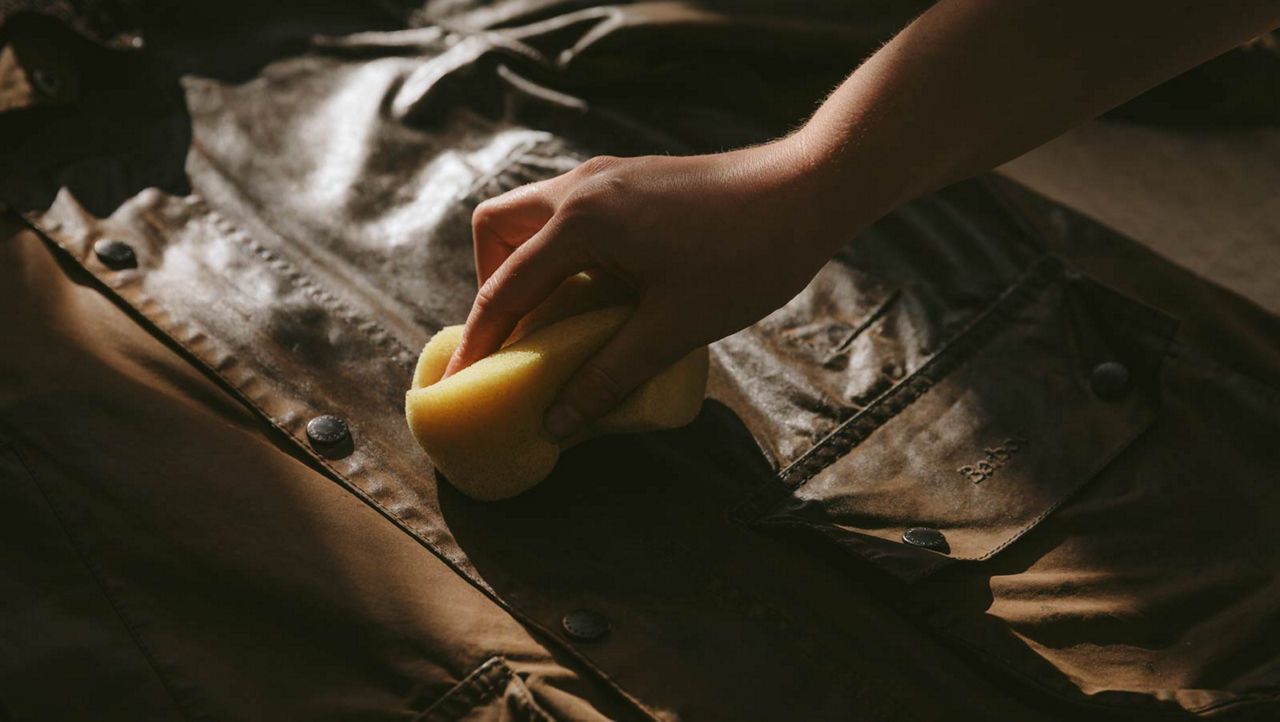 Pot of Barbour jacket wax in foreground, with someone waxing brown Barbour jacket in background with sponge