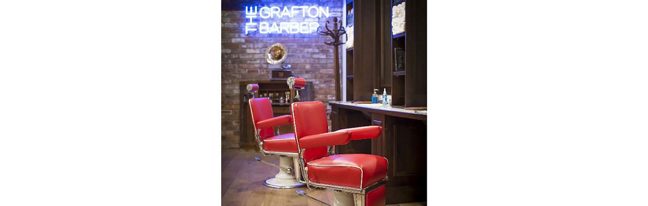 Two red barbers chairs with a neon 'The Grafton Barber' sign in the background