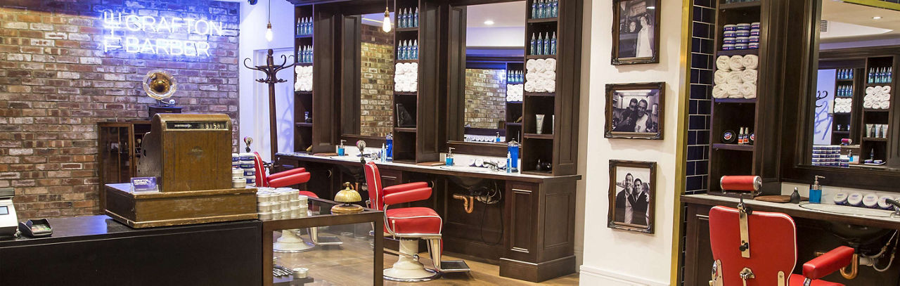 Barber shop interior with red chairs, vintage decor, mirrors, and shelves stocked with grooming products. Neon sign and framed photos adorn the walls.

