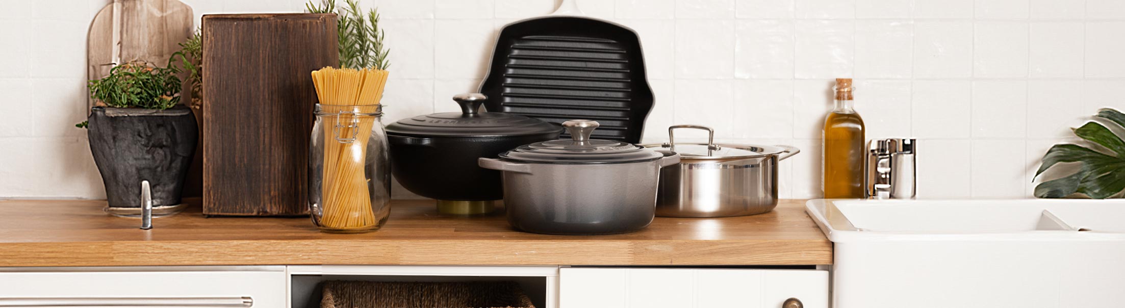 Kitchen counter with pots, a jar of pasta, cutting boards, and an oil bottle against a tiled wall.
