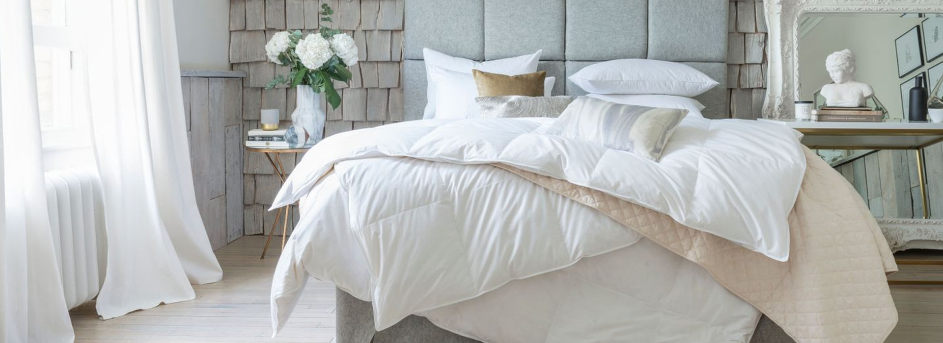 A cozy bedroom with a plush, gray-upholstered bed, white bedding, and decor elements like flowers and a bust on a side table.