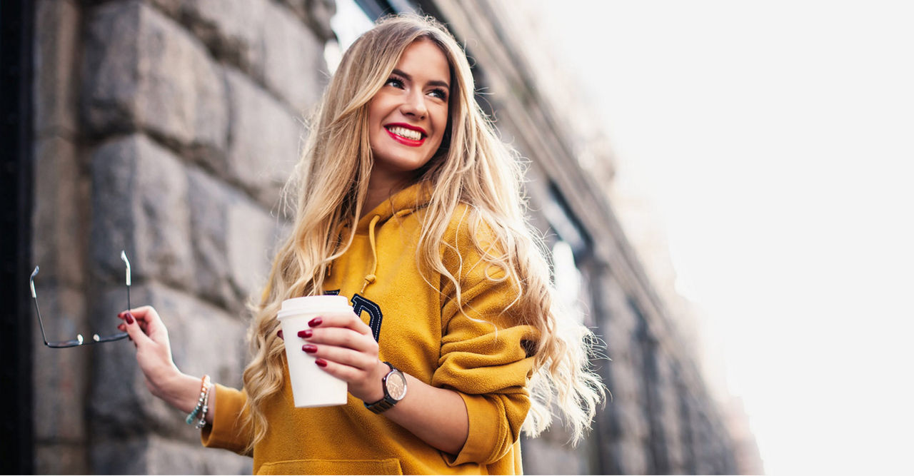 A woman in a yellow hoodie smiles, holding a coffee cup and sunglasses, standing beside a stone wall.

