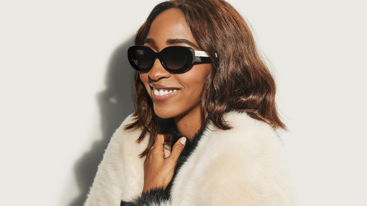 A woman wearing sunglasses and a faux fur coat smiles against a plain background.
