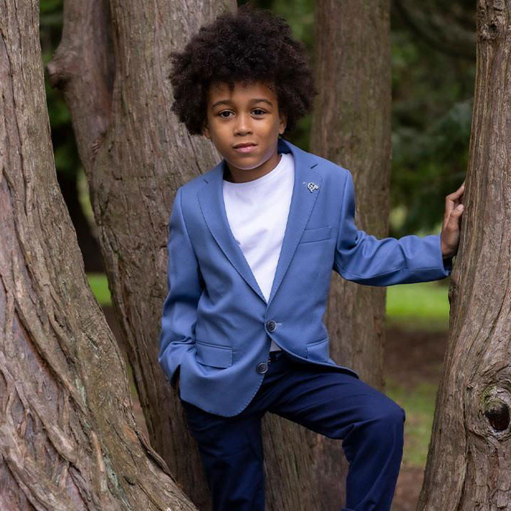 A child in a blue blazer and white shirt stands between tree trunks, set against a backdrop of greenery.