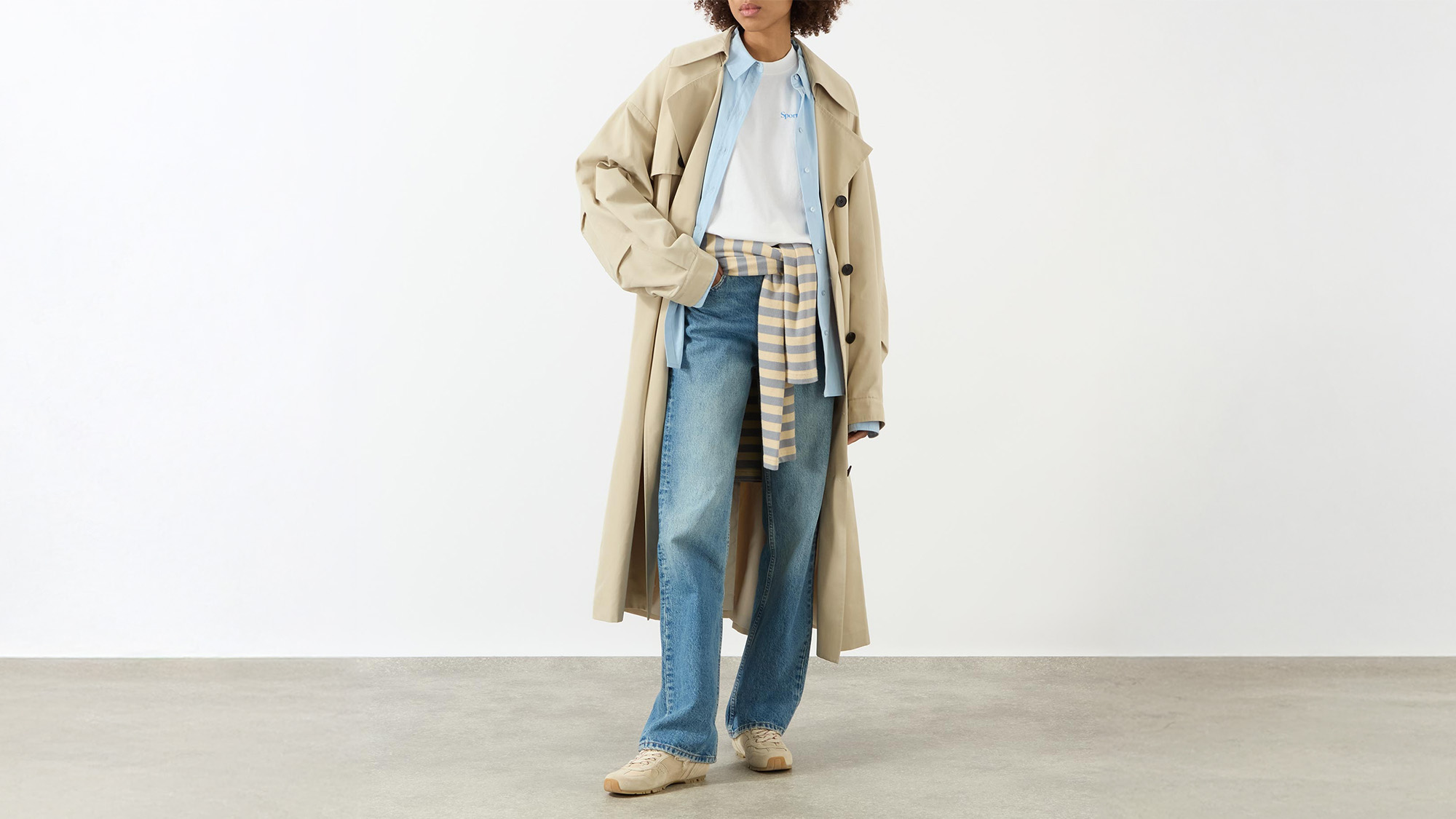 Person wearing a beige trench coat, blue jeans, sneakers, with a striped sweater around their waist and a light blue shirt underneath.
