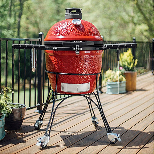 Red ceramic grill on a wooden deck, with potted plants and a black railing in the background.