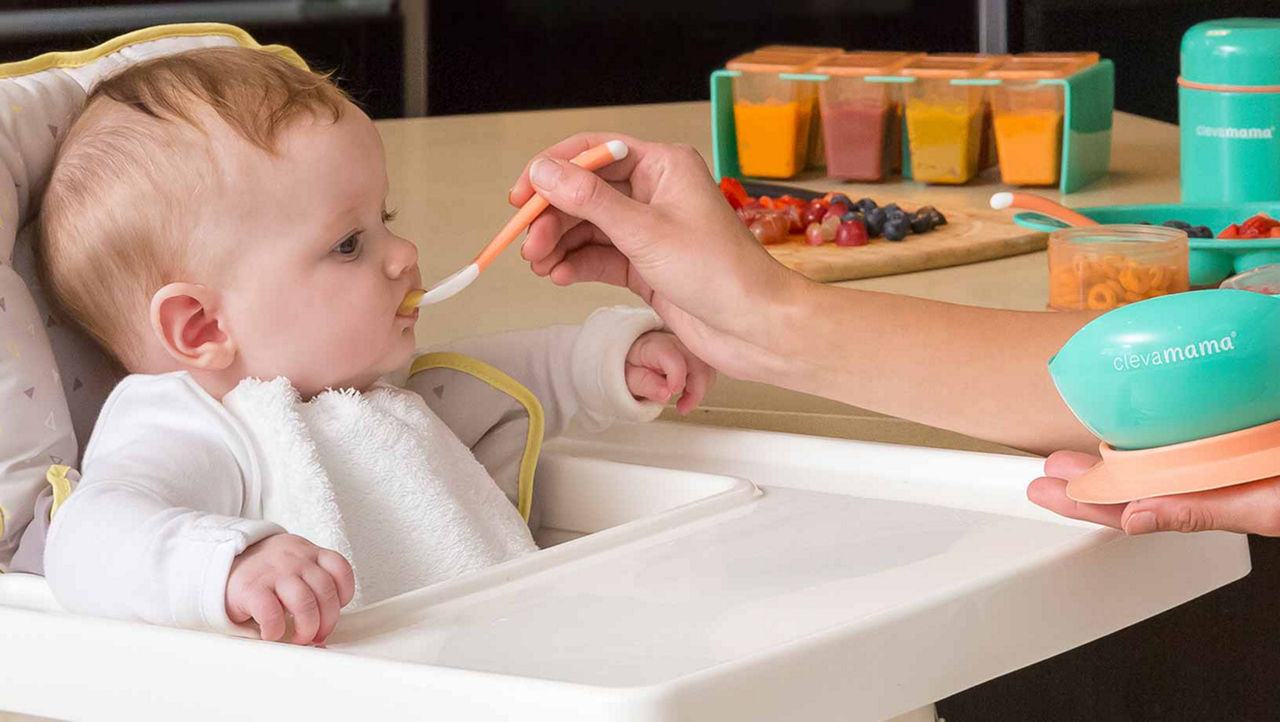 A baby sitting in a white high chair being fed with a white and orange spoon. The mother holding a blue and orange baby bowl.