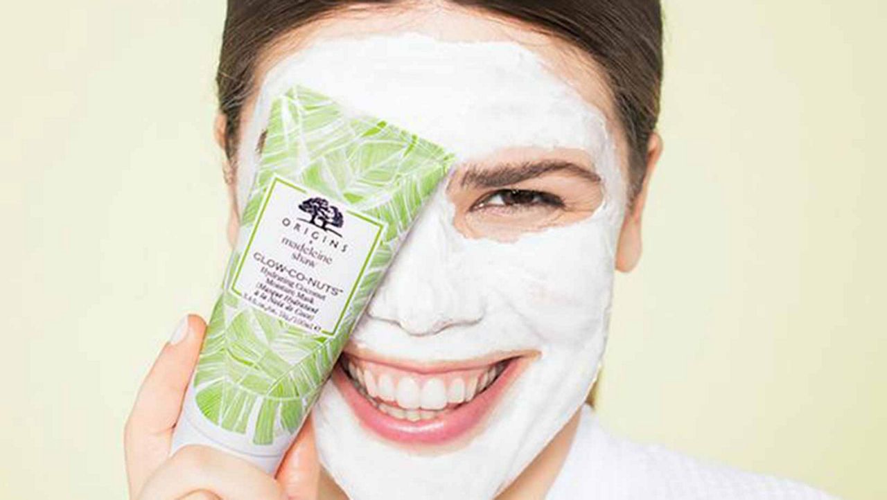 Model smiling with the origins Glow- Co-Nuts mask on her face, one eye covered with the product bottles she's holding up