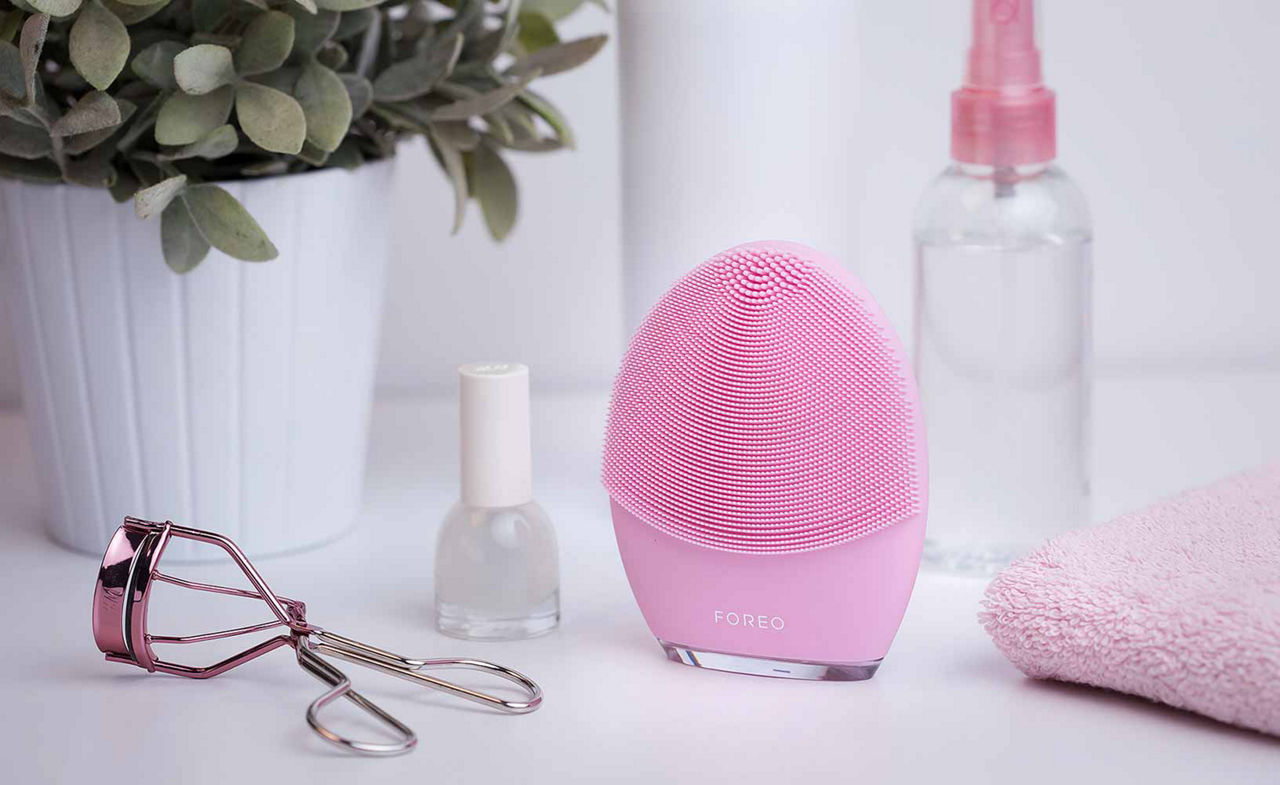 Foreo pink face messager on a white shelf surrounded by non discript bottles, a eyelash curler, pink towl and plant