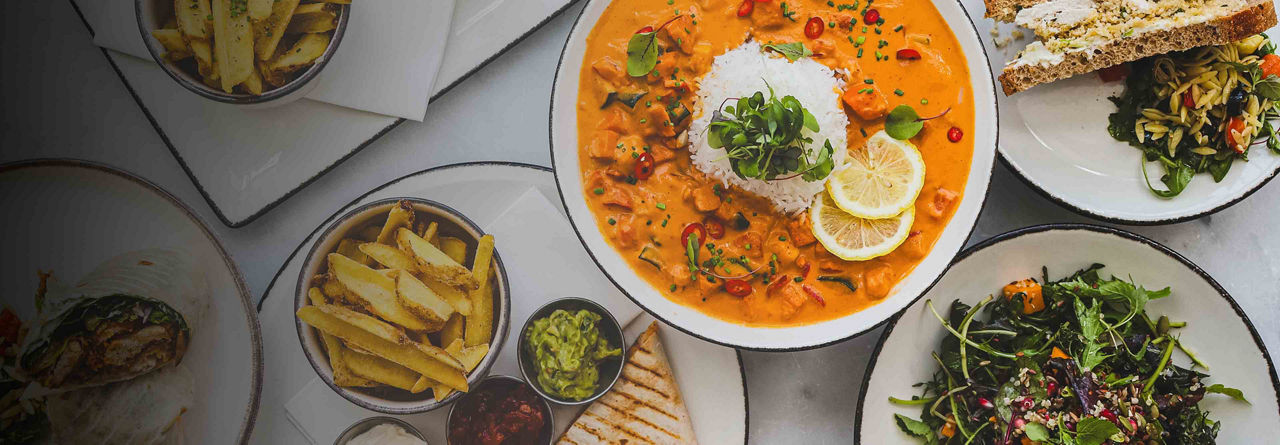 A variety of dishes including curry with rice, fries, avocado dip, sandwiches, and salads, arranged on a table.
