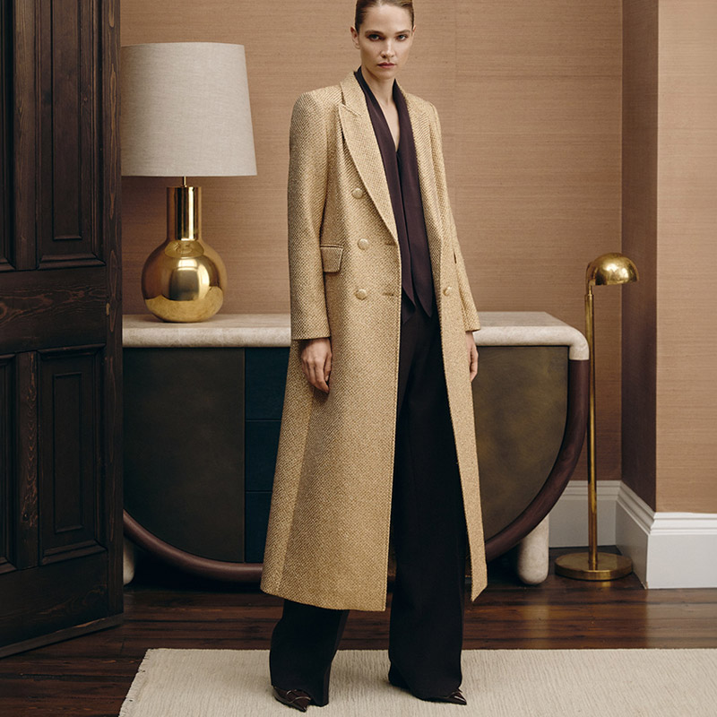 
A person stands in a room wearing a long beige coat over dark clothing. There's a lamp and a cabinet in the background.
