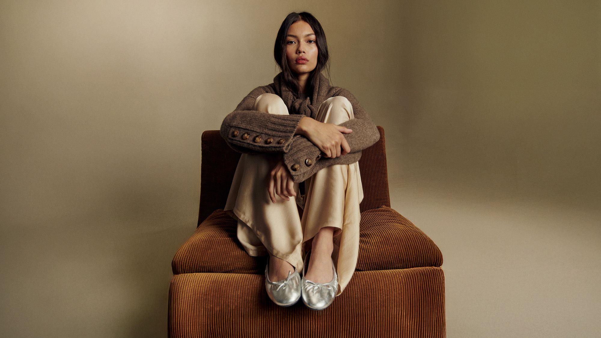 A person wearing a brown Sézane sweater, cream pants, and silver shoes sits on a chair. 