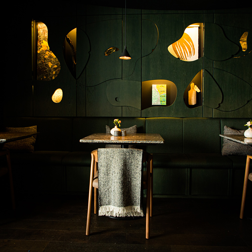 A dimly lit, modern dining area with abstract wall art, two tables set with vases and chairs, and a cozy ambiance.