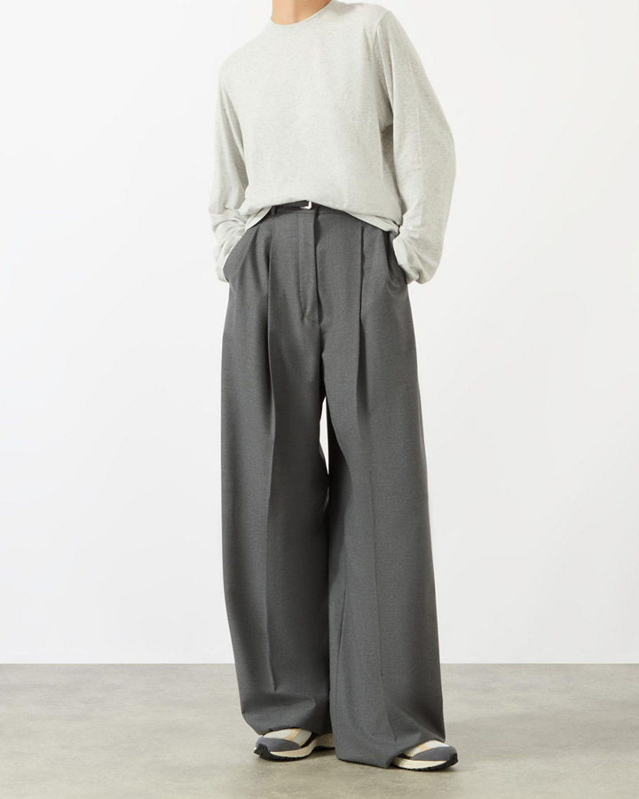 Person in a light sweater and wide-legged gray trousers with hands in pockets, wearing sandals.