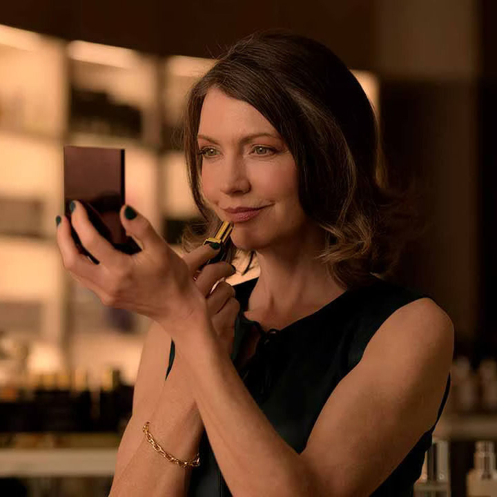 A woman applies lipstick while looking into a compact mirror in a softly lit room.