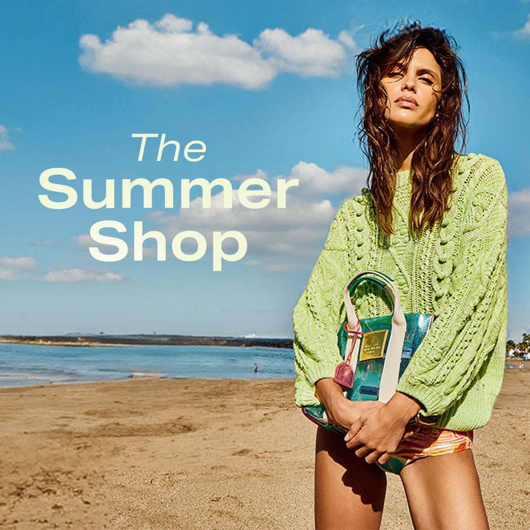 A woman in a green sweater holds a colorful bag on a beach. Text reads 'The Summer Shop.'