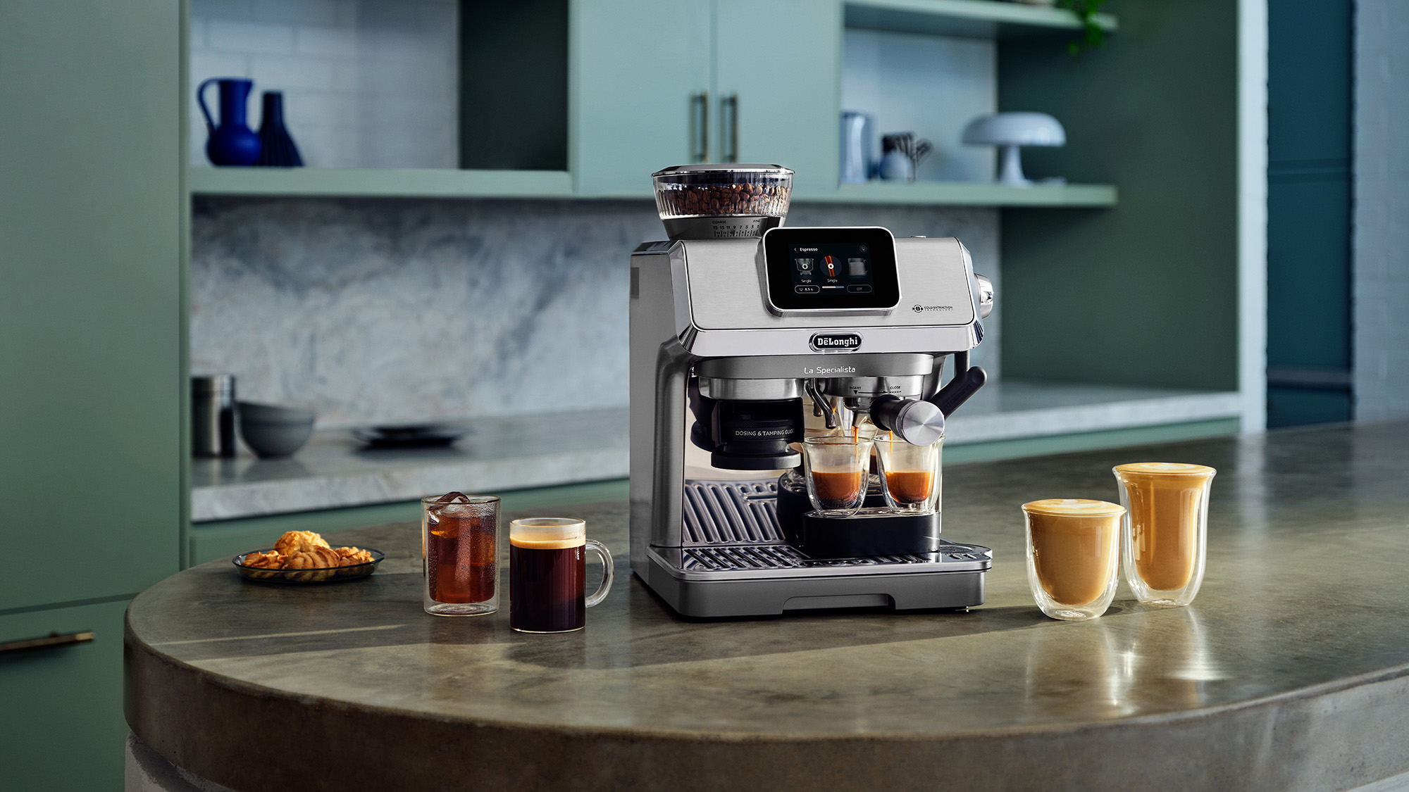 A modern espresso machine on a kitchen counter with a variety of coffee drinks and pastries nearby. The kitchen has green cabinetry.