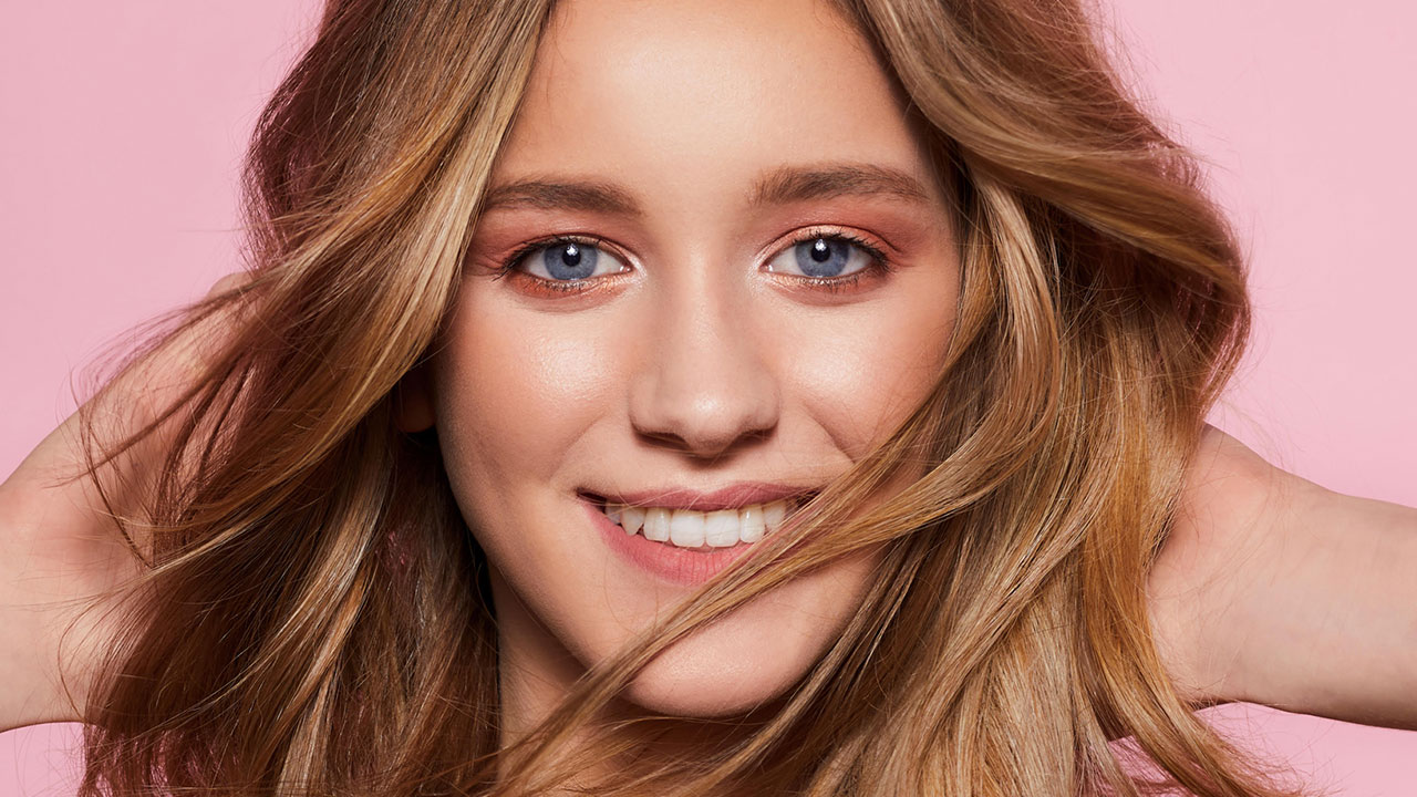 A smiling person with long brown hair holds their hair back, against a pink background.

